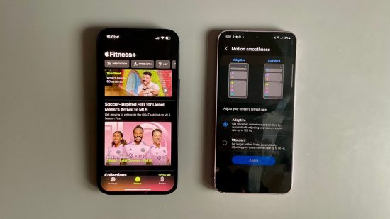 iPhone vs Android header showing an iPhone 14 in black and a Samsung Galaxy S23 in lavender. Both are face up, with the Apple Fitness app on the iPhone screen showing various marketing shots and features, while the Samsung is on a settings page for screen refresh rate variability.