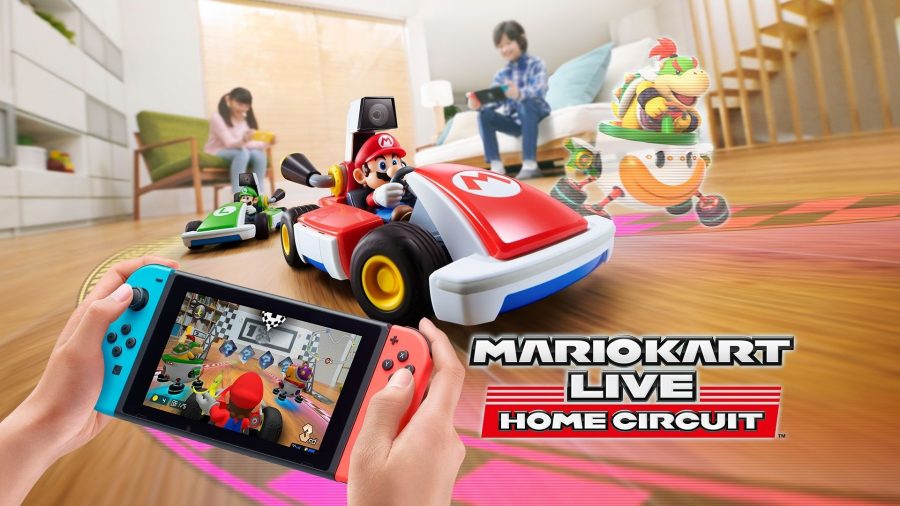 A person holds a Switch, showing the view from a camera on a real-life Mario kart, which is in from of them in a house, with kids playing behind it, from one of the many car games Mario Kart Live Home Circuit.